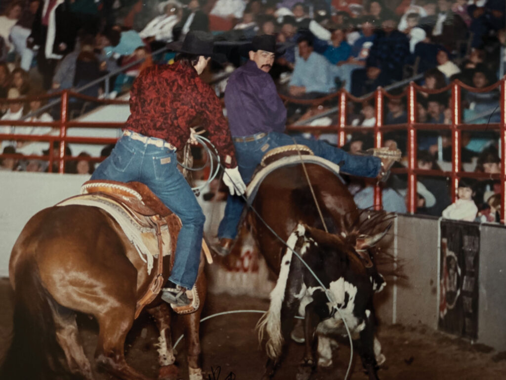 Two team ropers on sorrel horses work a roped steer in an indoor arena as a crowd watches from the stands
