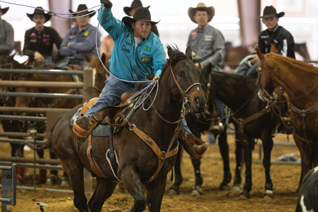 Header roper in a teal shirt and black cowboy hat swings his loop aboard a dark bay horse as mounted contestants watch from the arena fence