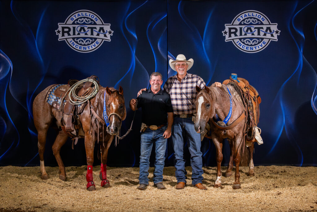 Two smiling men stand between saddled roan horses in front of a Riata Stallion Incentive backdrop