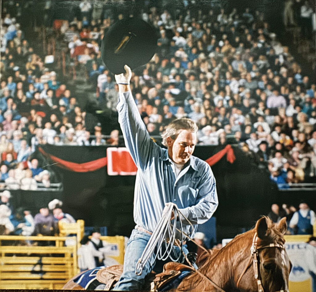 Cowboy on horseback raises his black hat to a packed arena crowd while holding a coiled rope in his other hand
