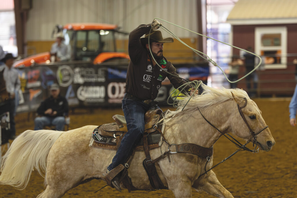 Header roper in a cowboy hat swings his loop overhead while galloping on a palomino horse across an indoor arena