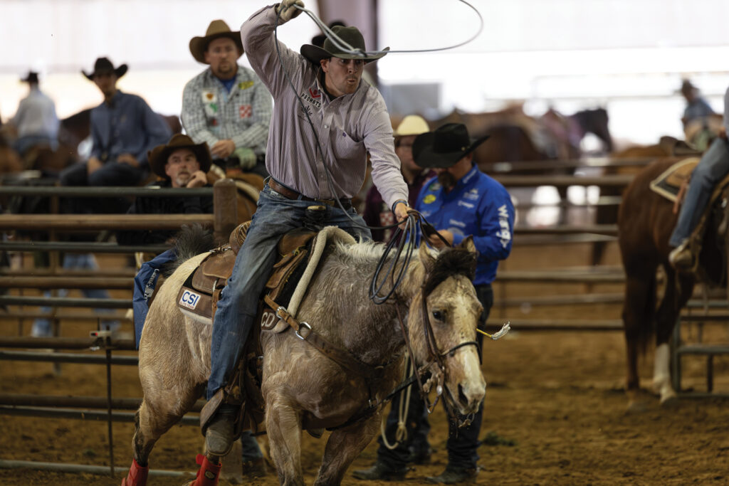 Header swinging his rope overhead aboard a gray horse as he rides out of the box in an indoor arena