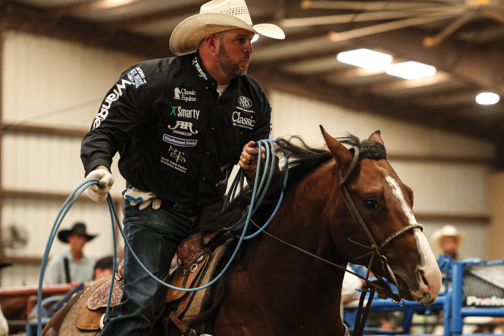 Team roper in a straw cowboy hat and sponsor-covered jacket swings his blue rope aboard a bay horse in an indoor arena