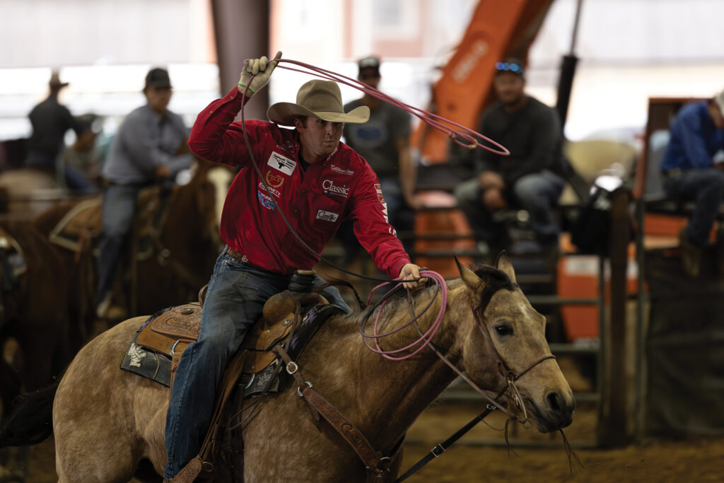 Cowboy in a red shirt and tan hat swings his rope overhead aboard a buckskin horse as spectators watch from the arena fence