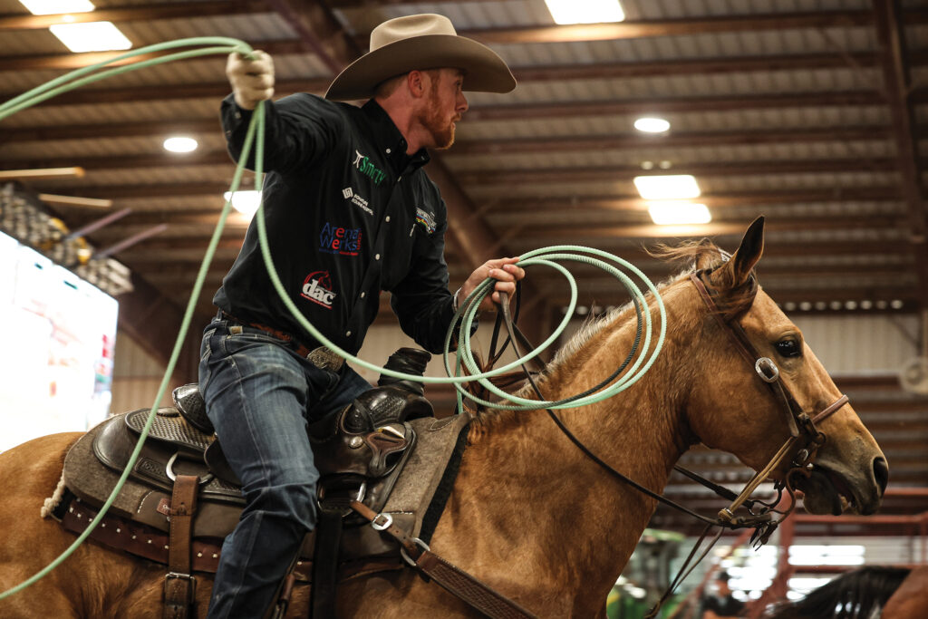 Team roper in a black shirt and tan cowboy hat swings his loop overhead aboard a palomino horse inside an indoor arena