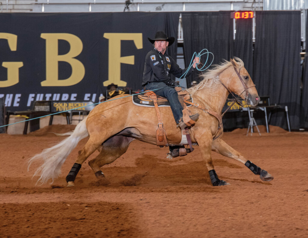 Header roper in black hat and vest swings his loop as his palomino horse slides to a stop in the arena dirt
