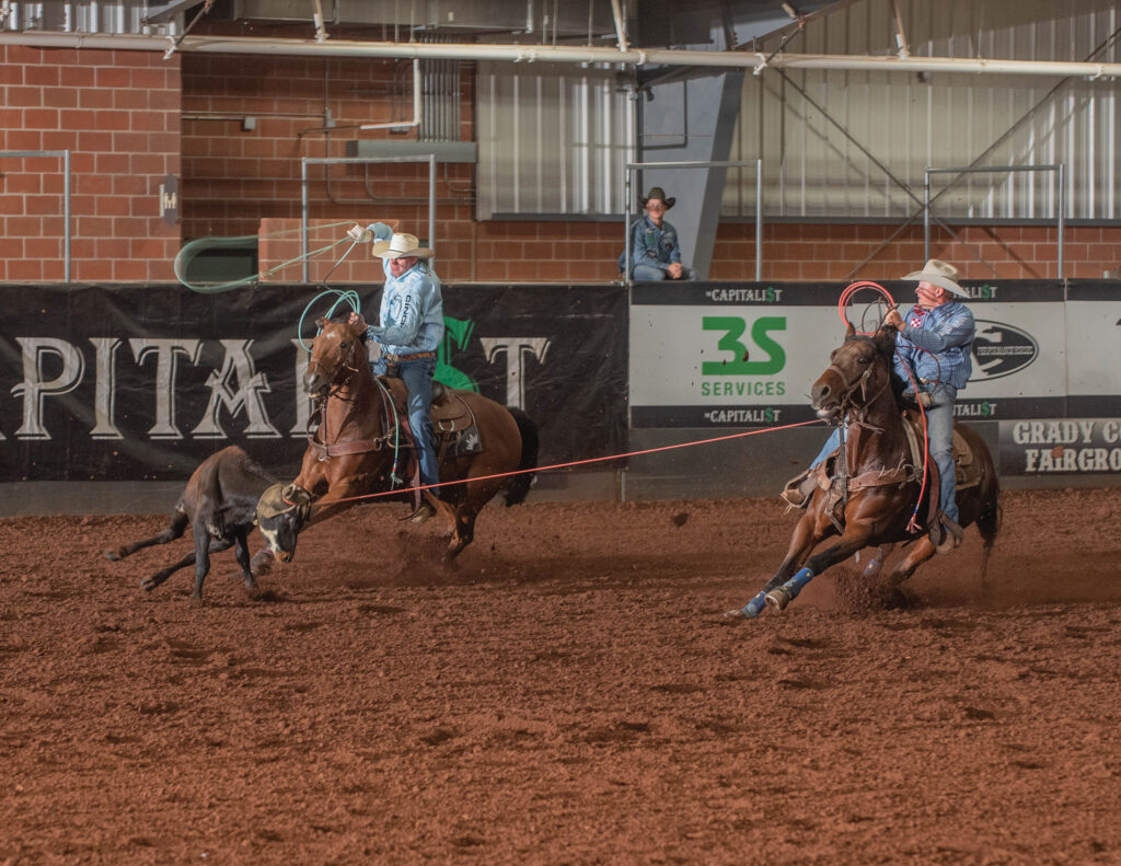 Two team ropers on horseback chase a steer across an indoor arena, the header swinging his loop as the heeler's rope stretches tight