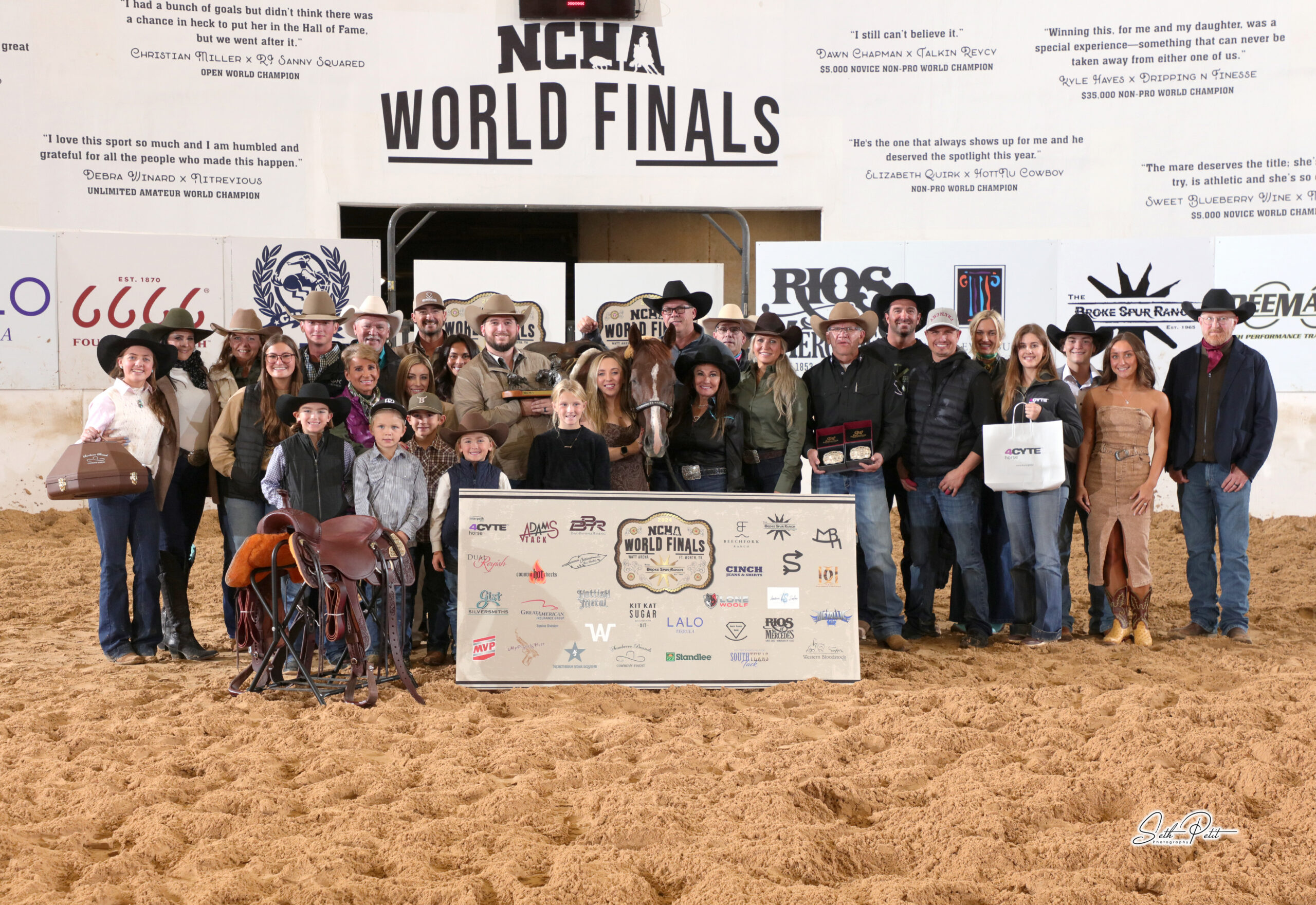 Large group of family, friends and sponsors gathered with a horse, trophy saddles, buckles and sponsor banner in the arena at the NCHA World Finals