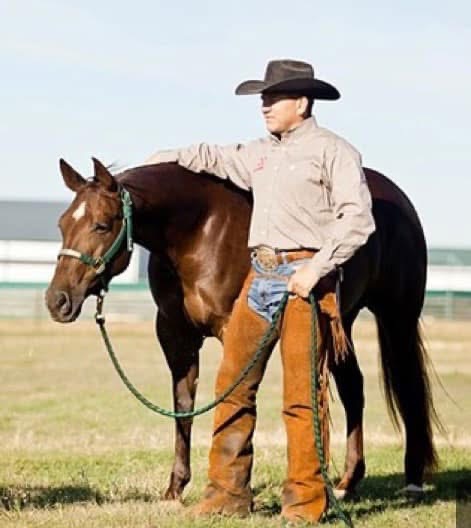 Cowboy in a black felt hat and leather chaps stands beside a sorrel horse in a green rope halter on grass
