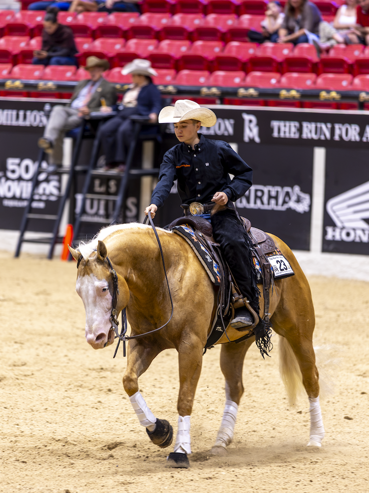 Cowboy in black shirt and tan hat rides a sorrel horse across an arena during a Western riding event at an indoor rodeo venue