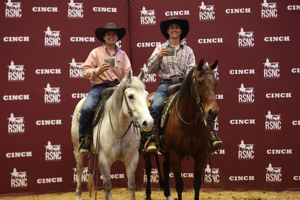 Two smiling riders in cowboy hats hold up trophy belt buckles aboard a gray and a bay horse before a Cinch RSNC backdrop