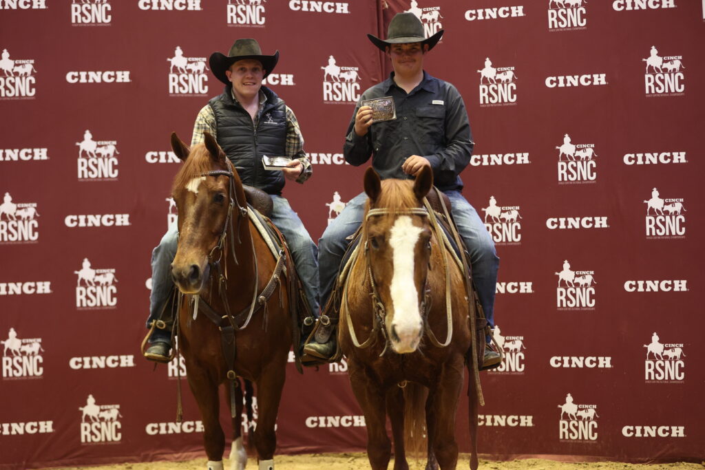 Two cowboys on sorrel horses hold championship belt buckles in front of a Cinch RSNC backdrop