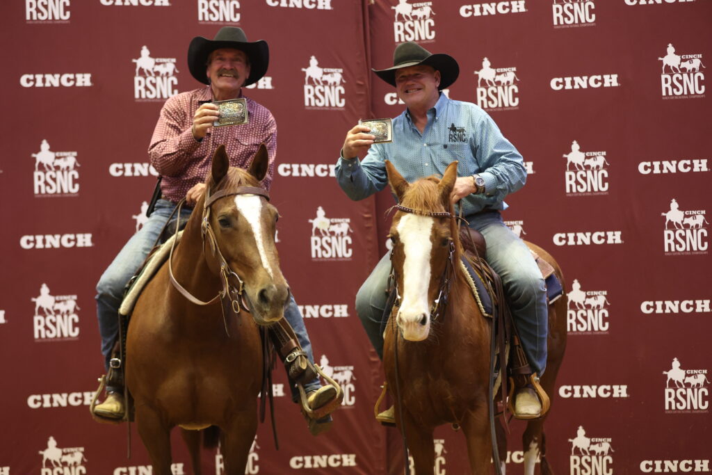 Two riders in cowboy hats hold up championship belt buckles while mounted on sorrel horses before a Cinch RSNC backdrop