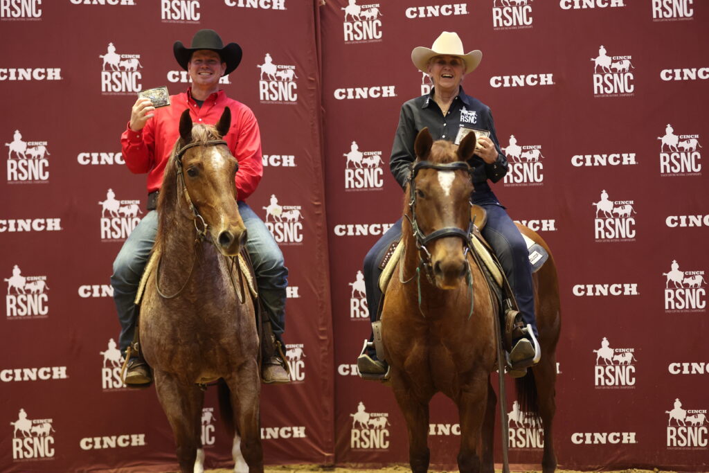 Two mounted riders hold up championship belt buckles in front of a Cinch RSNC backdrop