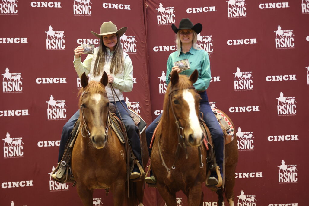 Two women in cowboy hats hold up champion belt buckles while mounted on sorrel horses before a Cinch RSNC backdrop