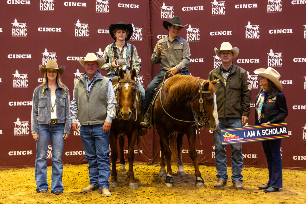 Two young riders holding buckles sit horseback beside sponsors and an "I Am A Scholar" sign at a Cinch RSNC awards presentation