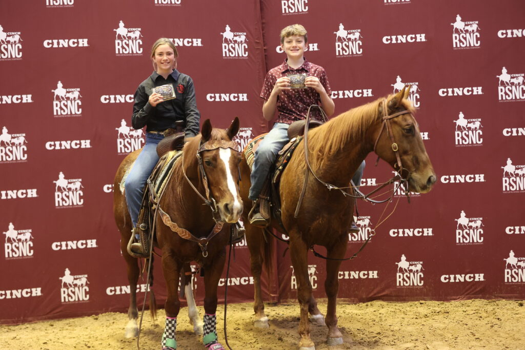 Two young riders on sorrel horses hold up championship belt buckles in front of a Cinch RSNC backdrop