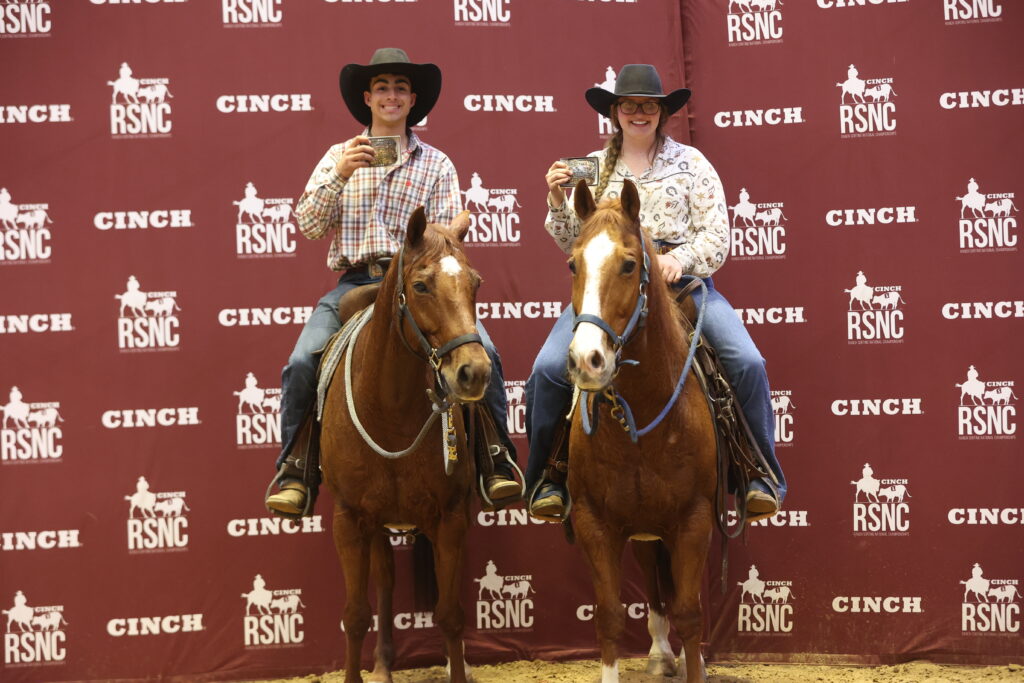 Two mounted riders hold up championship belt buckles in front of a Cinch RSNC backdrop