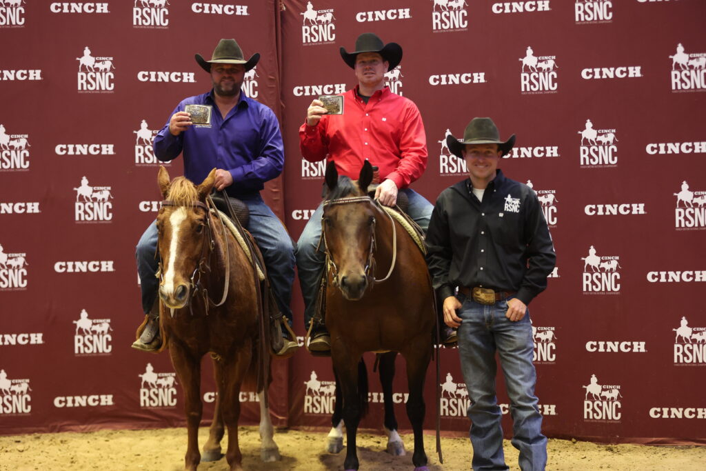 Two mounted riders hold up championship belt buckles beside a man standing, in front of a Cinch RSNC backdrop