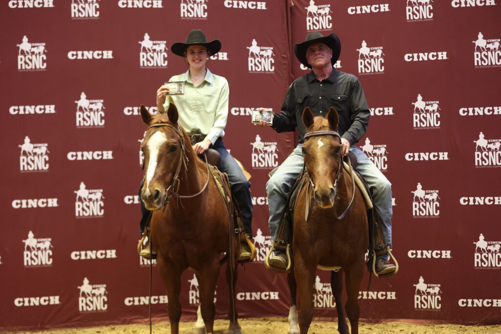Two riders on sorrel horses hold up belt buckles in front of a Cinch RSNC backdrop after a ranch sorting win