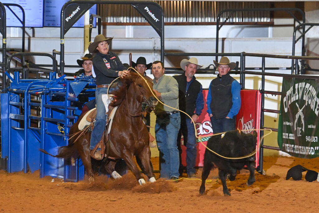 Breakaway roper swings her loop from a sorrel horse as a black calf runs ahead, with spectators watching from the arena fence