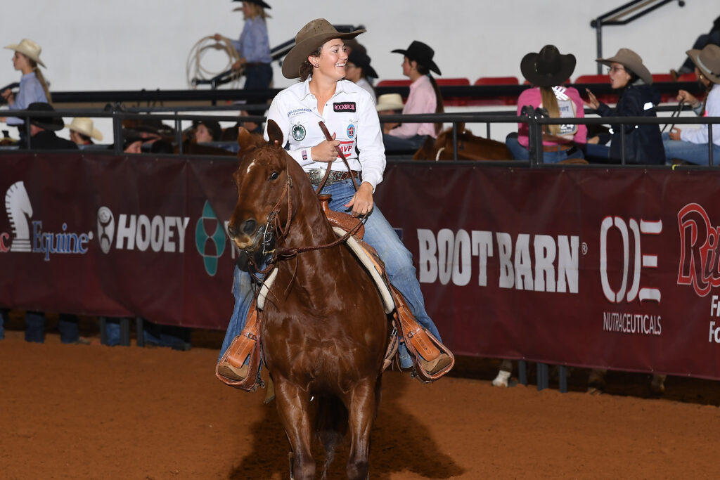 Smiling cowgirl in a tan cowboy hat and white shirt rides a sorrel horse past arena banners after her run