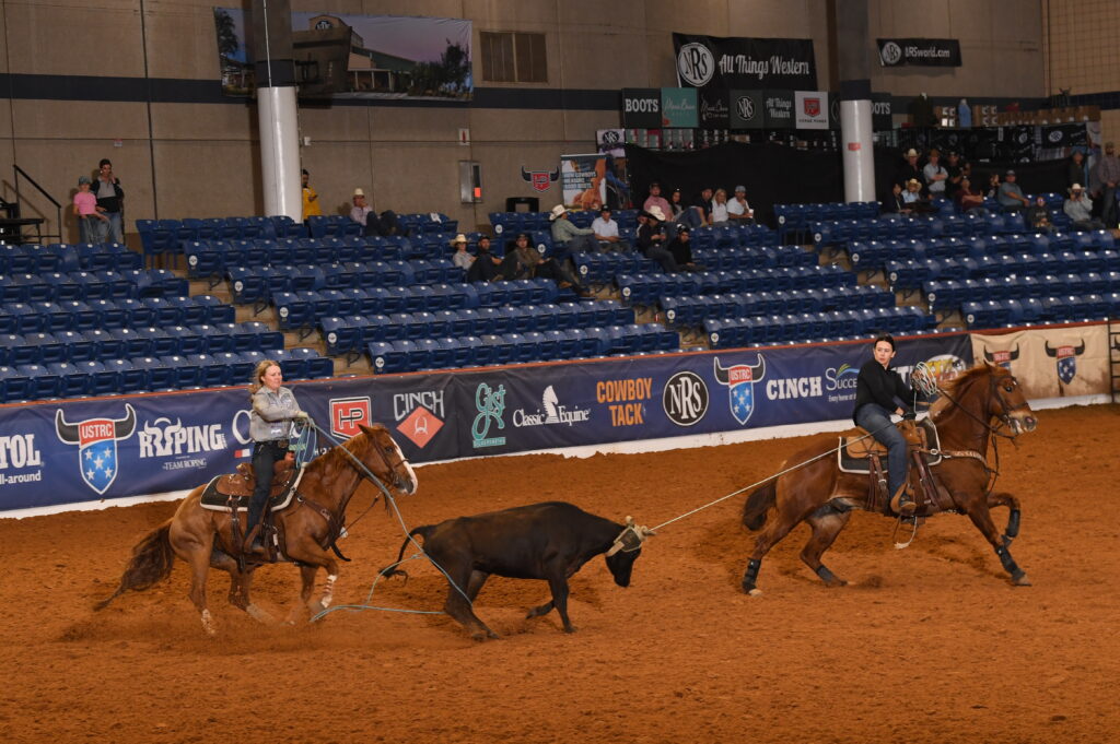 Two team ropers on horseback work a steer in a professional indoor arena with sponsor signage and sparse spectator seating