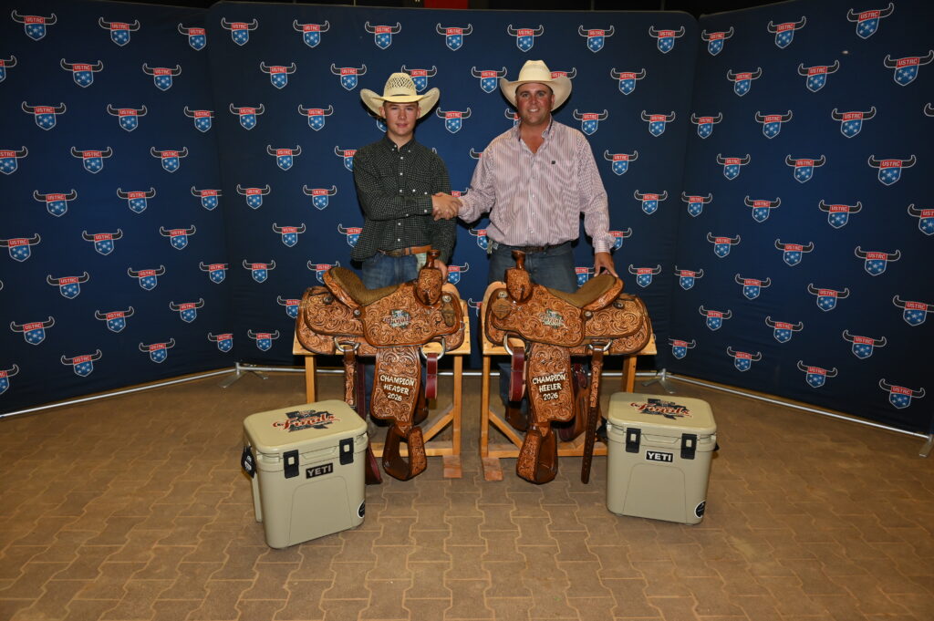 Two cowboys in western attire shake hands while posing with ornate leather saddles and YETI coolers against a branded backdrop