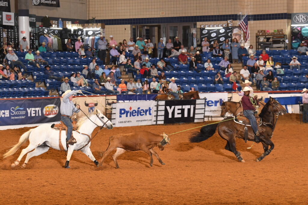 Team ropers work a steer during competition at an indoor rodeo arena with YETI and Priefert sponsorship signage visible