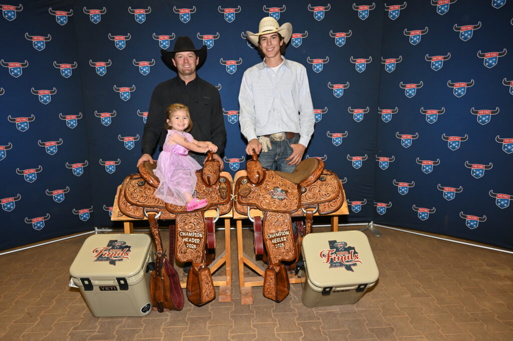 Two men and a child pose with ornate leather saddles at a rodeo finals event backdrop