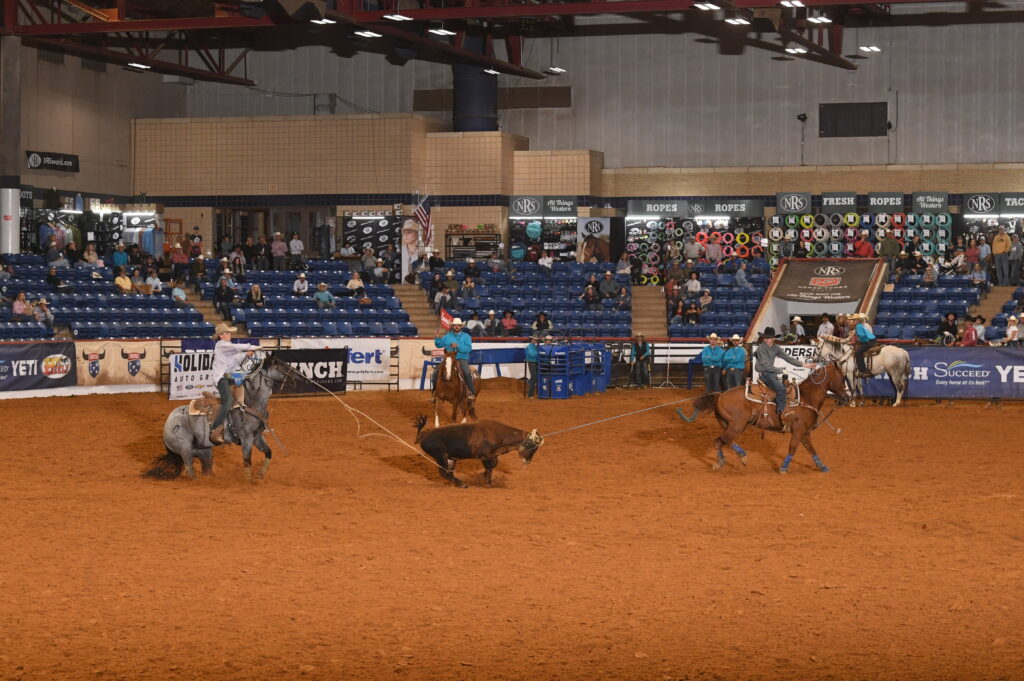 Three team ropers work a steer in the arena during an indoor rodeo event with spectators in the stands