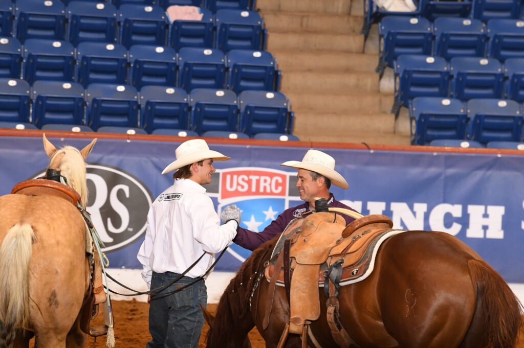 Two cowboys in western attire converse while holding horses in a rodeo arena with USTRC signage visible