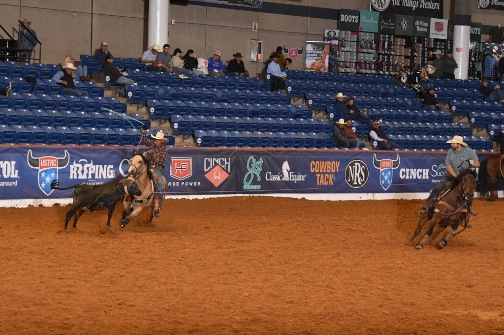 Two team ropers on horseback work cattle in a covered arena during a rodeo competition, with spectators in the stands and sponsor signage visible along the barrier
