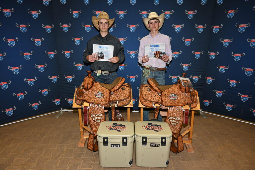 Two young men in cowboy hats display awards and certificates at a rodeo event, standing behind ornate leather saddles and YETI coolers branded with rodeo logos