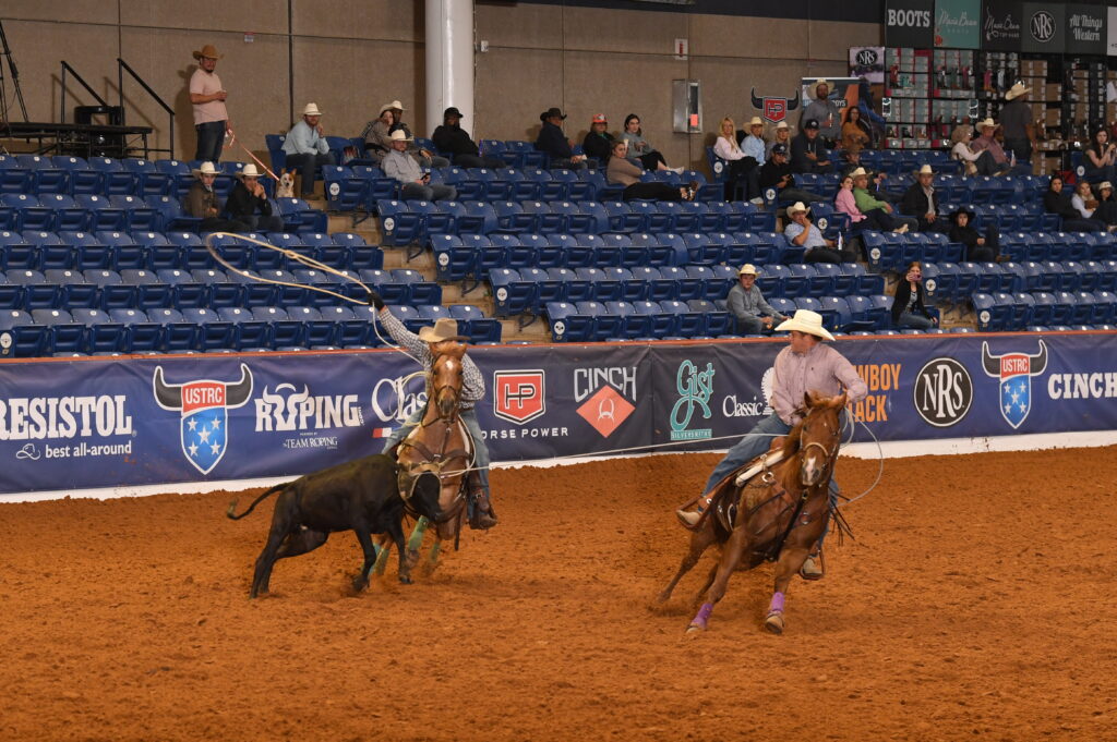 Two ropers on horseback work a steer during team roping at an indoor rodeo arena with sponsor signage and spectators in blue seats