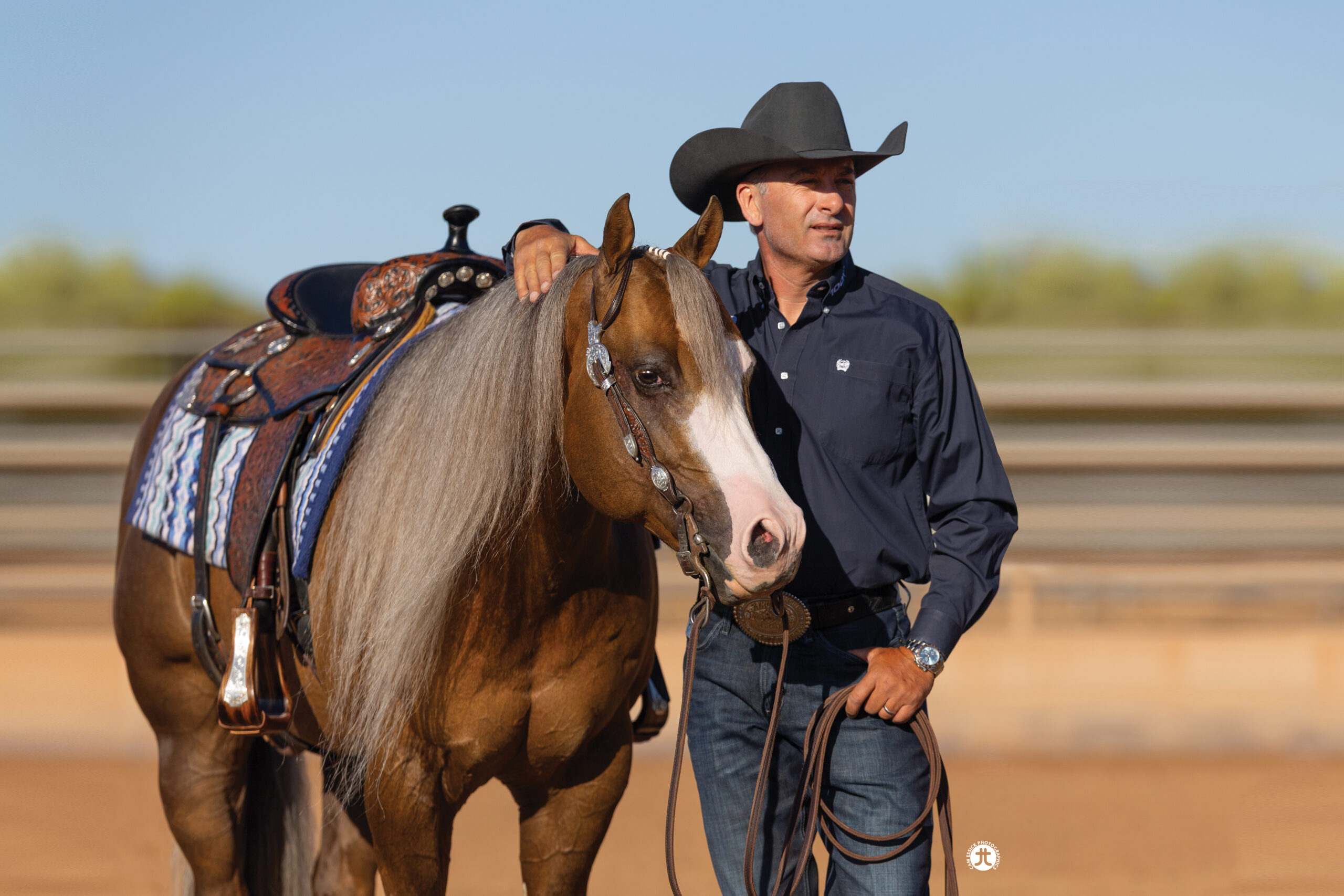 Man in black cowboy hat and navy shirt leads a saddled palomino horse with flaxen mane across a dirt arena
