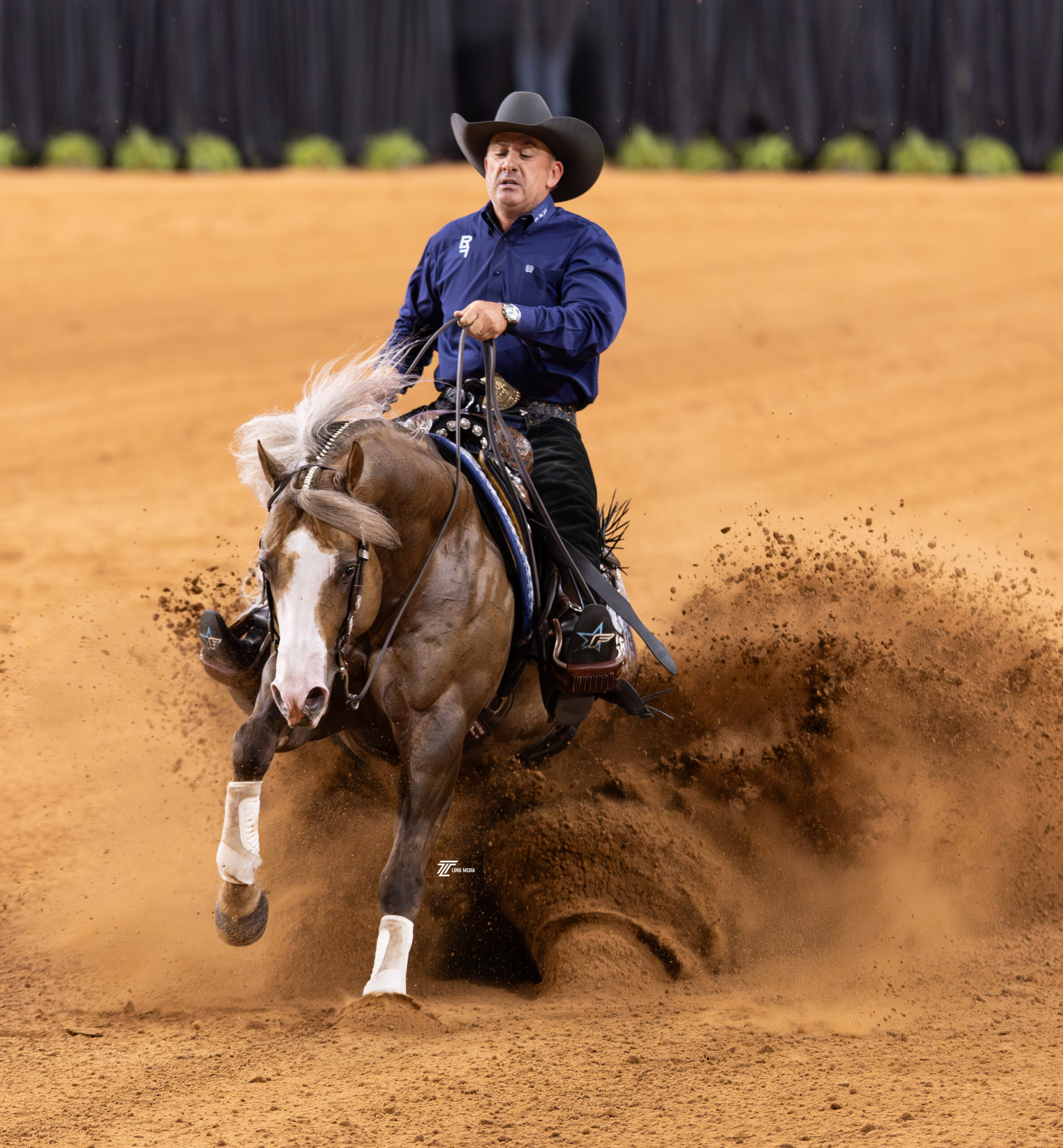 Reiner in a navy shirt and black cowboy hat slides a palomino horse to a stop, spraying dirt across the arena