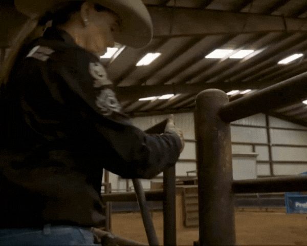 Cowboy in hat and jacket grips wooden arena rail inside indoor rodeo facility