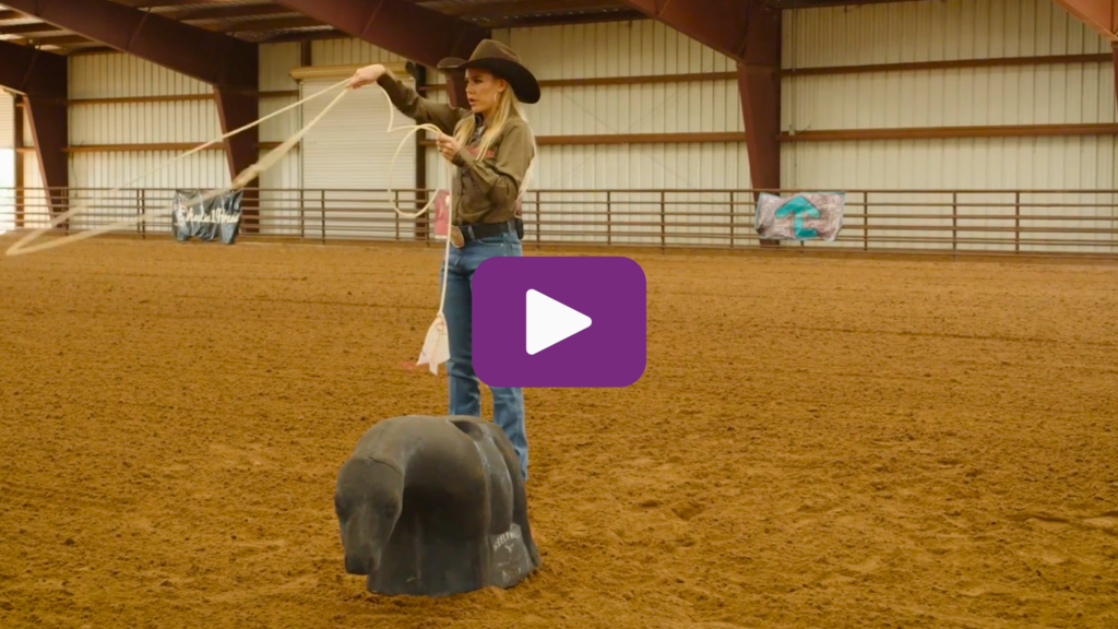 Woman in a cowboy hat swinging a rope toward a roping dummy steer in a covered arena