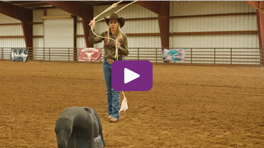 Breakaway roper in a cowboy hat swings her loop overhead as she practices on a roping dummy in an indoor arena