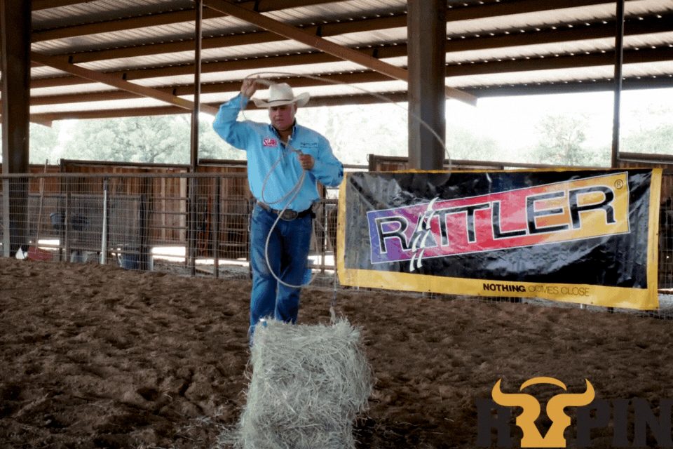 Cowboy in blue shirt and hat holding rope in sand arena with Rattler banner and cattle pen visible