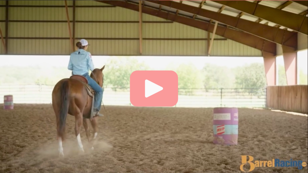 Rider in a light blue shirt and ball cap works a sorrel horse around barrels in a covered indoor arena