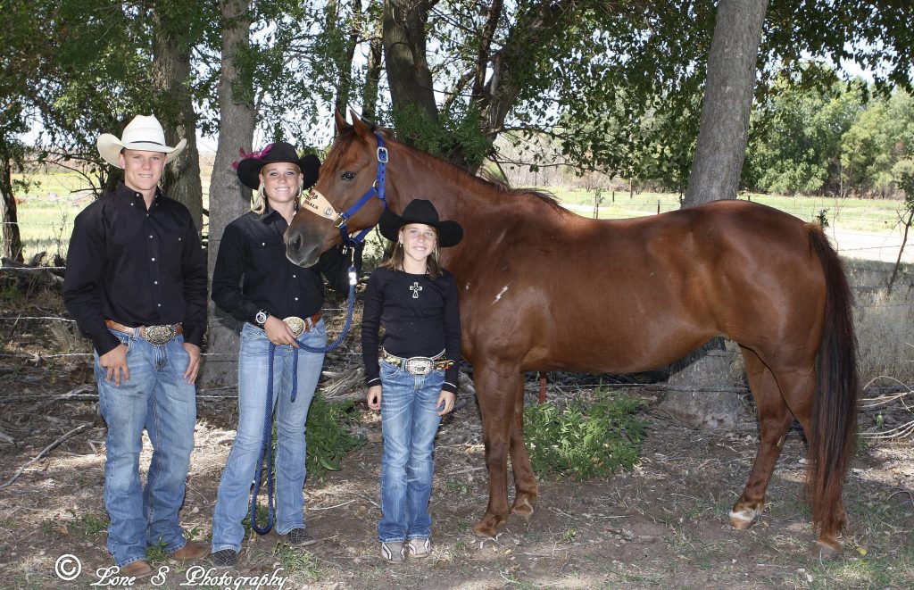 Three young rodeo competitors in black shirts and cowboy hats stand with a chestnut horse in a blue halter under shade trees