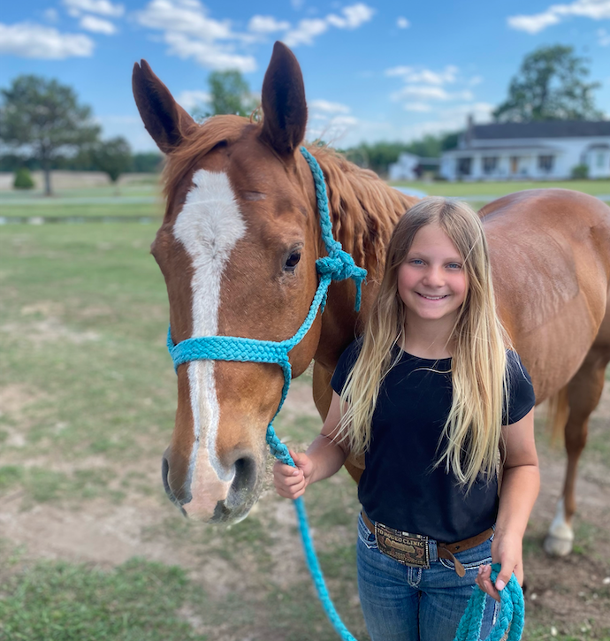 Smiling young girl in a black tee and trophy buckle belt stands beside a chestnut horse in a teal rope halter and lead