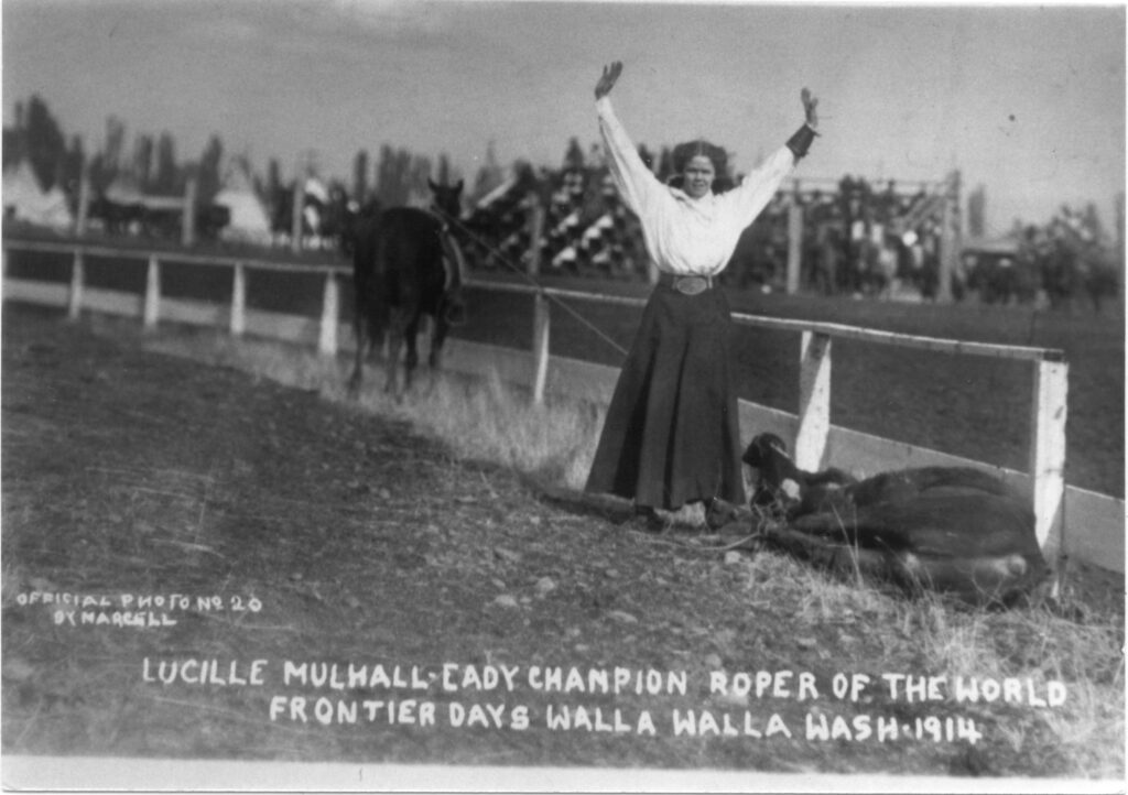 Historic 1914 rodeo photo of a woman in long skirt raising both arms beside a roped, tied-down steer as her horse holds the rope taut