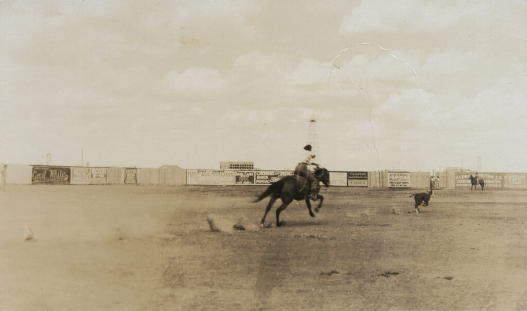 Vintage sepia rodeo arena shot of a rider galloping after a loose calf past a fence lined with painted advertisements
