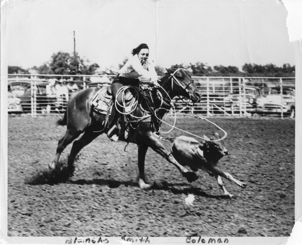 Vintage black-and-white rodeo photo of a woman roping a calf from horseback in a dirt arena as her loop sails out