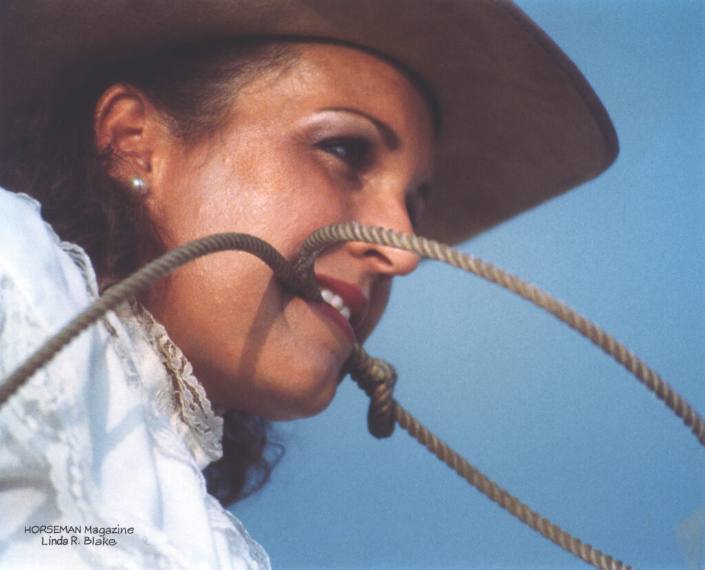 Smiling cowgirl in a wide-brimmed hat holds a lariat rope in her teeth against a clear blue sky