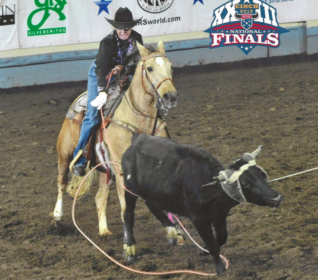 Header roper in black hat and jacket on a buckskin horse holds her rope as a roped black steer is stretched at the USTRC National Finals