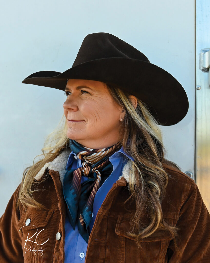 Woman in a black cowboy hat, brown corduroy jacket and silk wild rag scarf looks off to the side at a rodeo